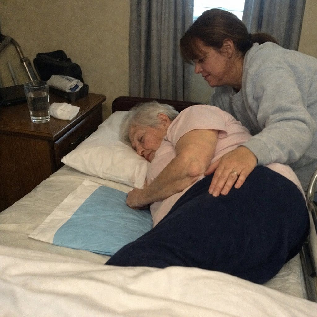 A middle-aged caregiver leans over an elderly woman lying on a care bed with a waterproof pad, providing hands-on hygiene assistance in a home bedroom