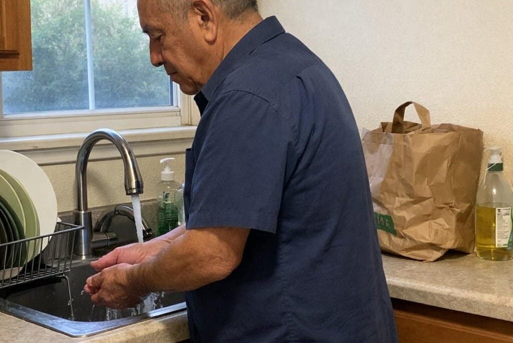 An older man standing in profile at a kitchen sink with both hands held open and relaxed under running water, photographed from across the kitchen near the table