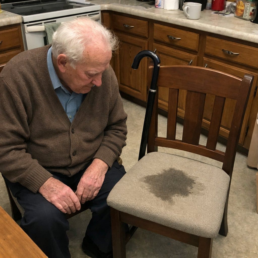 An elderly man sitting at a table looking down at a damp spot on the cushion of a nearby chair
