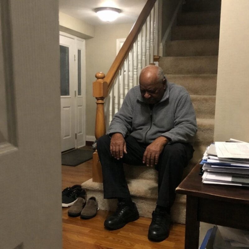 An older man sits on the lower steps of a home staircase with his hands on his knees, looking down at the floor, resting