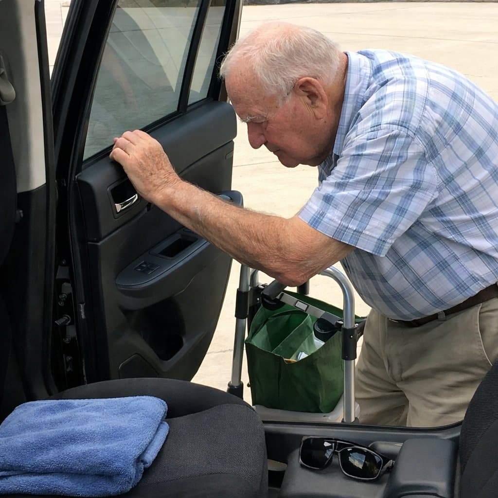 An older man with a walker parked behind him pressing the back of his hand against a hot rear car window to test the temperature before anyone gets in