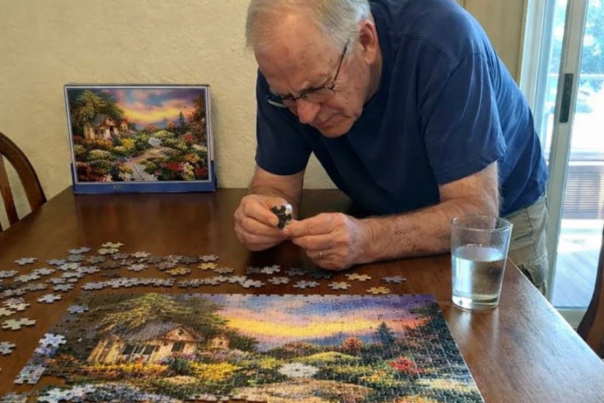 An older man stands over a dining table fitting a large puzzle piece into a half-finished jigsaw, squinting as he lines it up, the puzzle box propped against the wall behind the table
