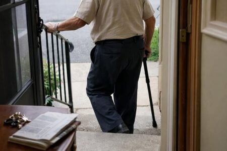 An older man using a cane descending two outdoor concrete steps while reaching for a single iron post handrail mounted at the edge of the stoop