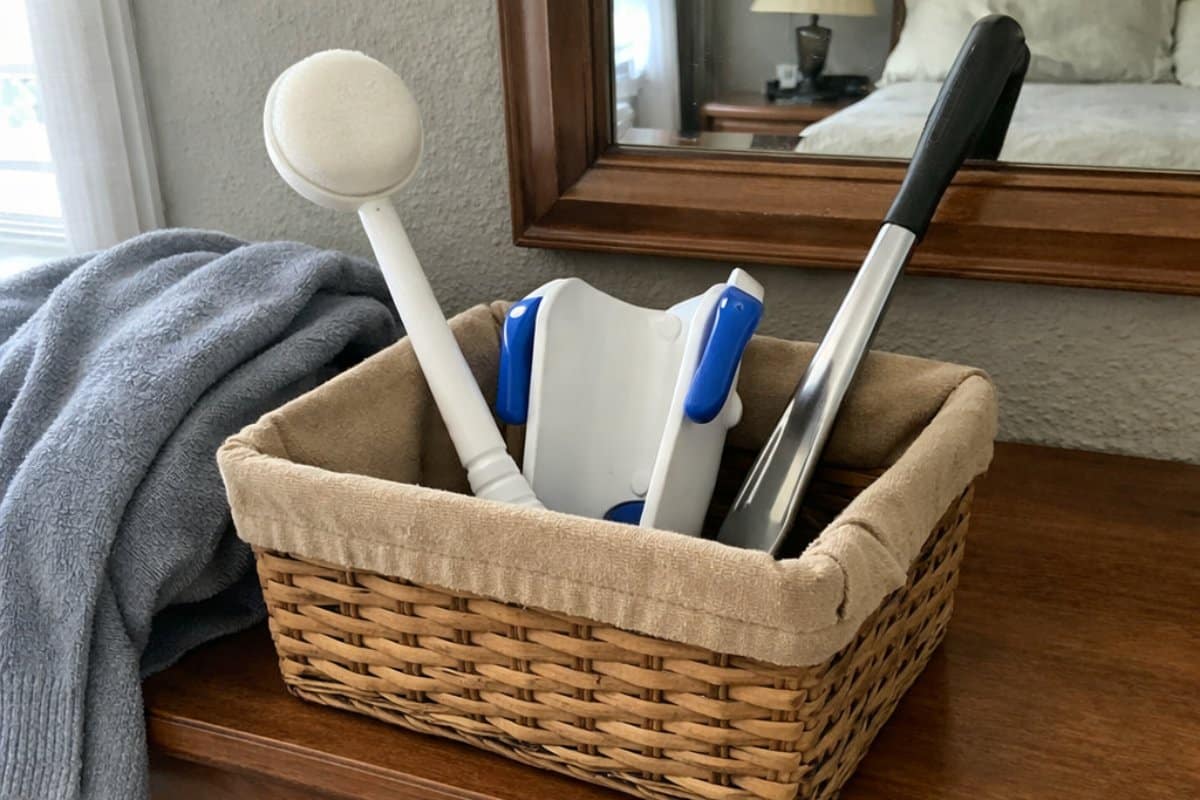 A basket on a bedroom dresser holding a long-handled lotion applicator, sock aid, and shoe horn, with slippers on the floor below