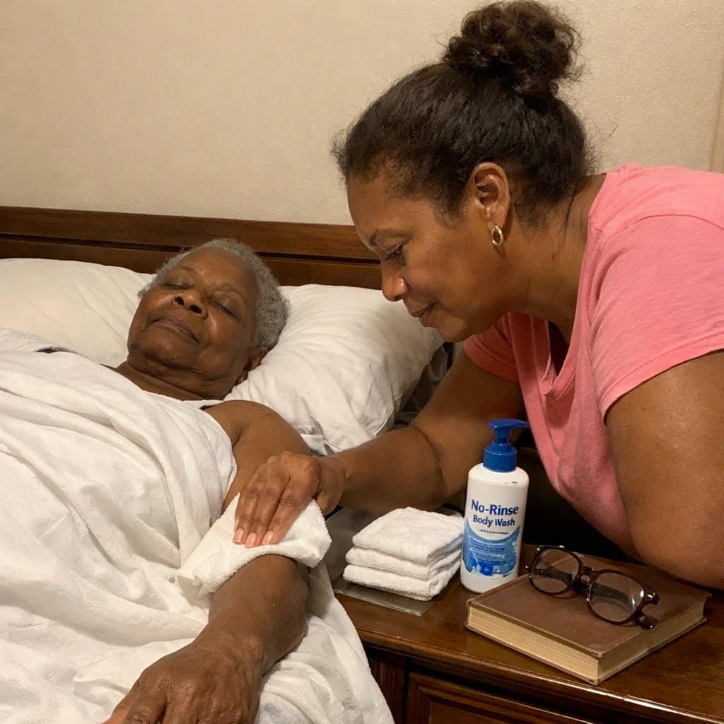 An older woman lies in bed under a sheet while her daughter wipes her forearm with a cloth, bathing supplies on the nightstand beside them