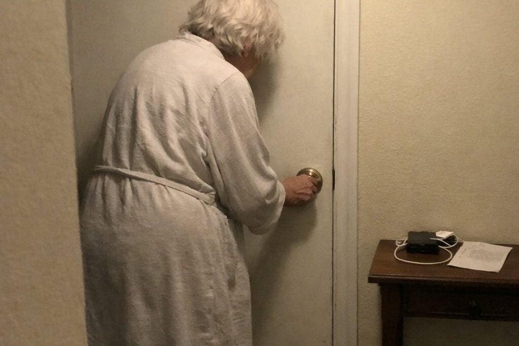 An older woman in a robe gripping a round door knob in a dim hallway at night, wrist twisted, bracing against the door