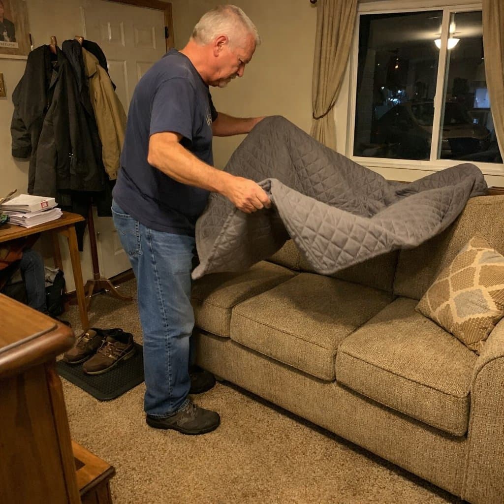 An older man standing at the end of a couch shaking out a large waterproof furniture protector before laying it down