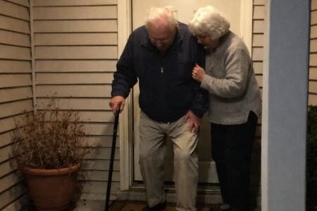 An older couple on a front porch step at night, one using a cane, both looking down carefully as they descend with the porch light on above them