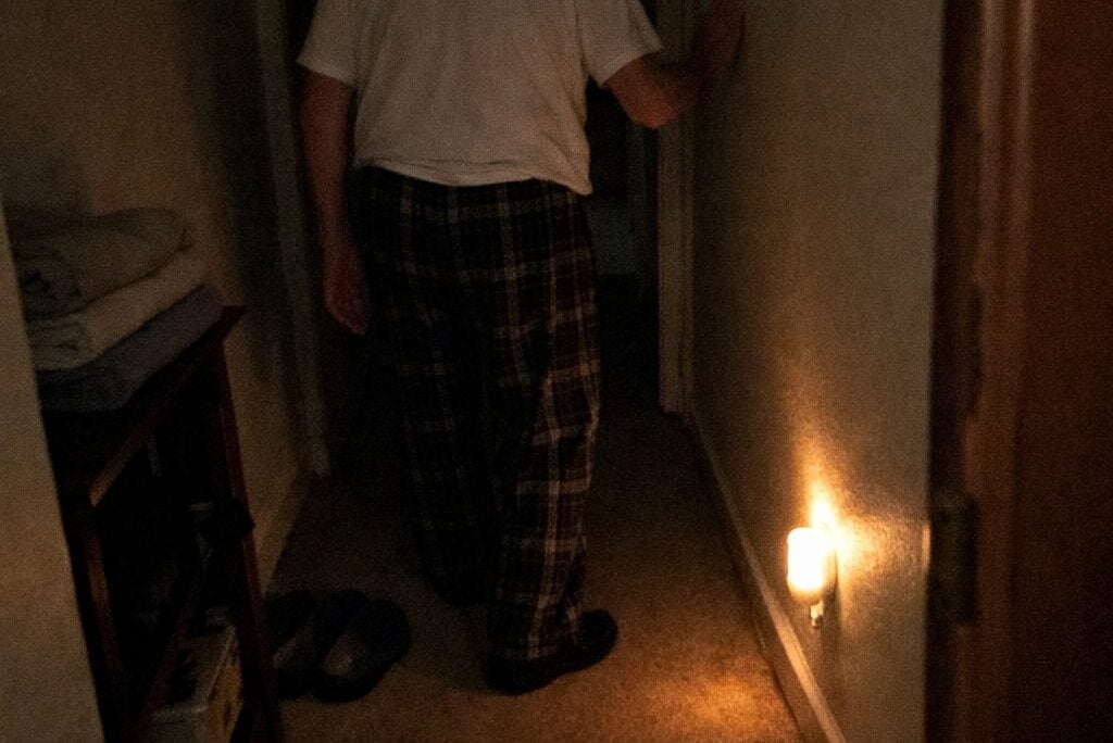 An older man walking slowly down a dark hallway toward a bathroom, with only a small plug-in night light illuminating a small section of the floor
