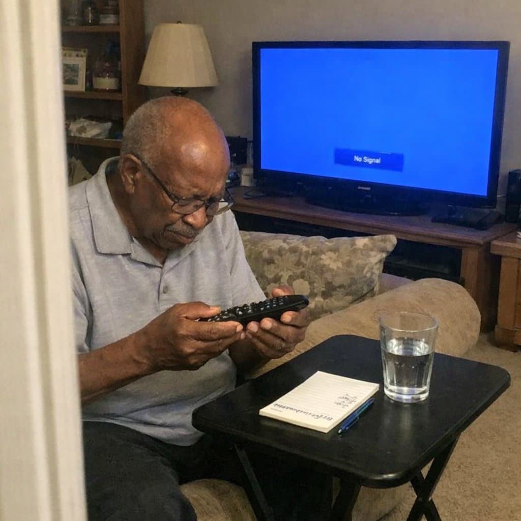 An older man sitting on a sofa turning a standard TV remote over in his hands and squinting at the buttons, with the TV showing a blue input screen in the background