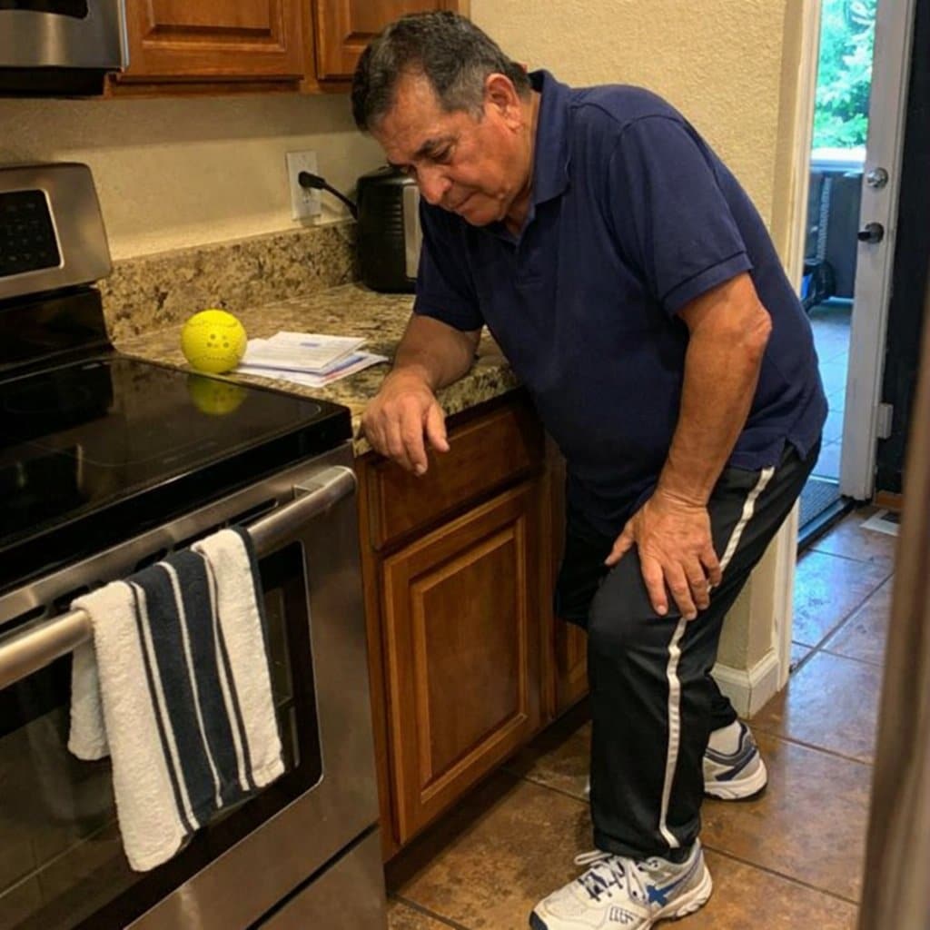 An older man standing at a kitchen counter pressing his hand against his knee, still wearing court shoes, with a pickleball on the counter and mail nearby