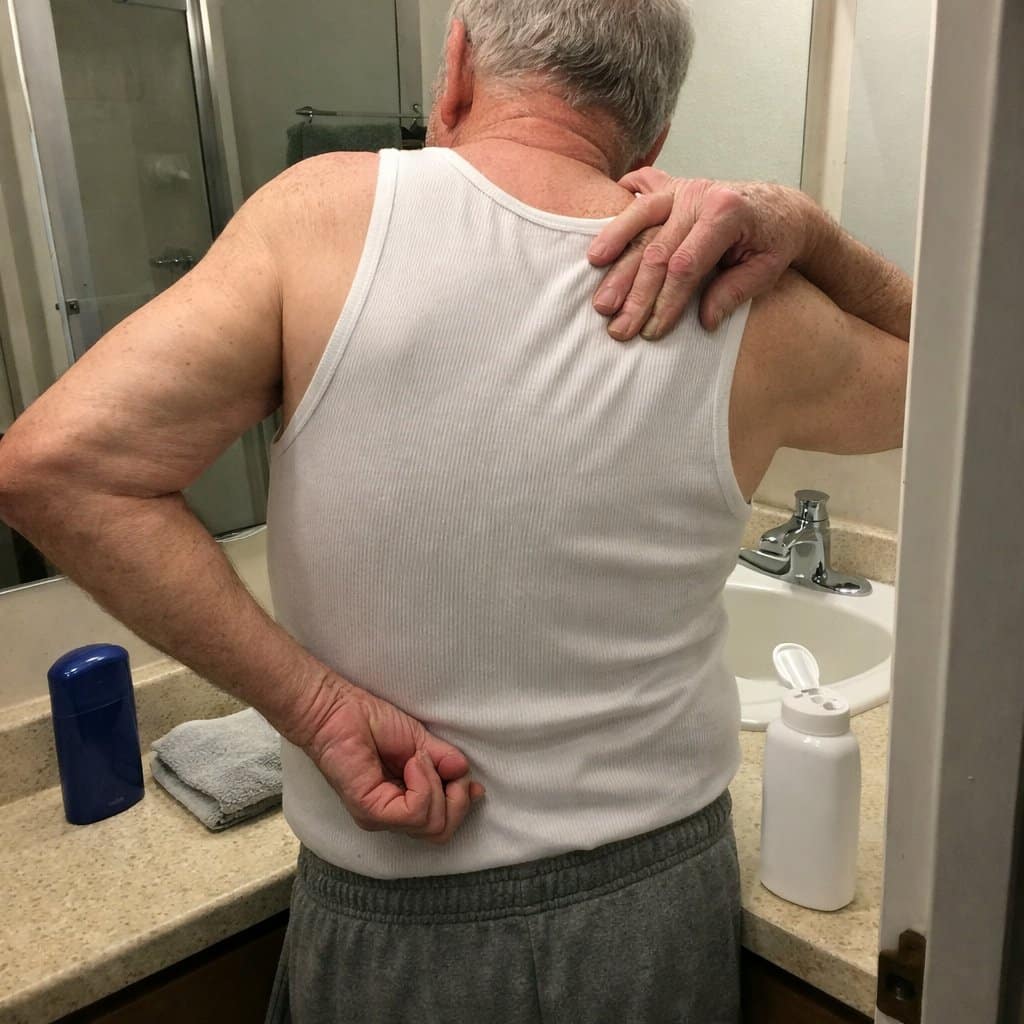 An older man in an undershirt twisting his arm behind his back to apply lotion, standing at a bathroom counter with personal care items