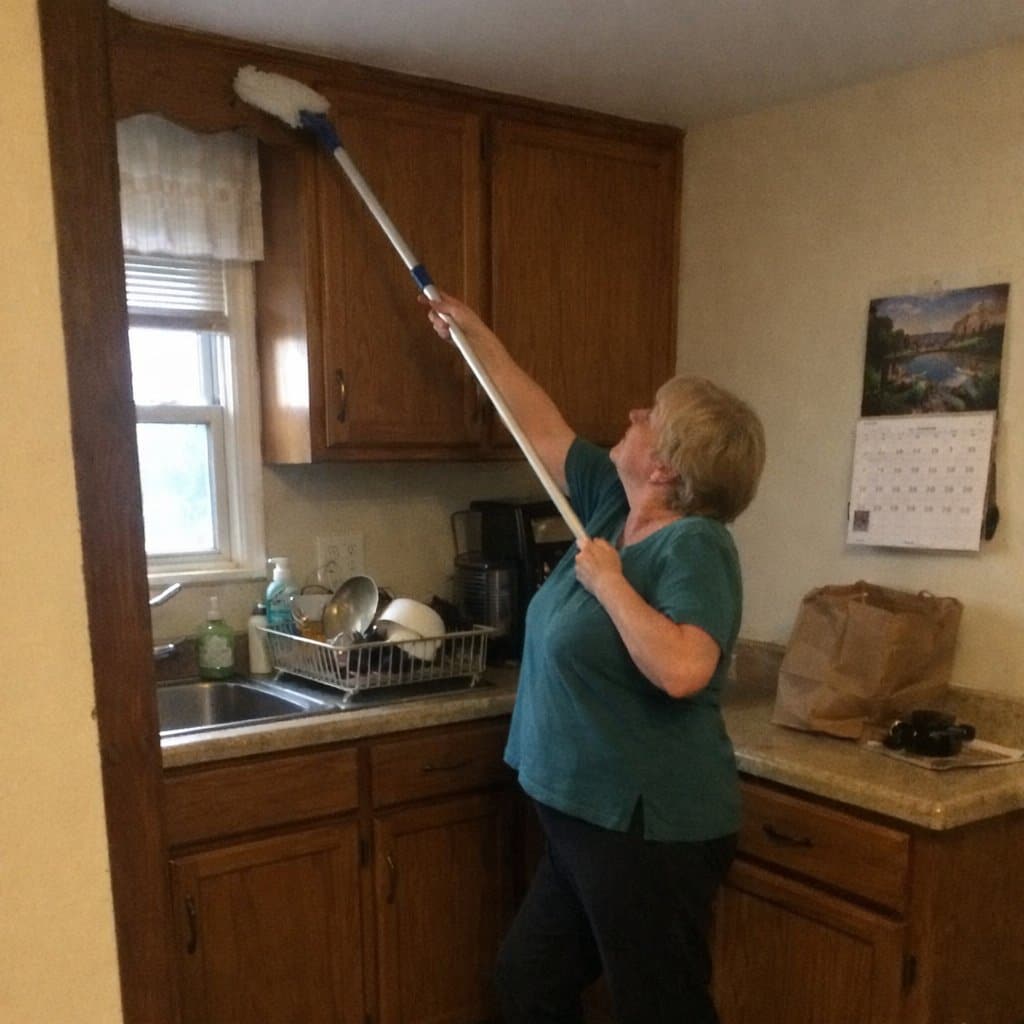 An older woman standing in a kitchen, using a long-handled tool to clean the top of an upper cabinet, stretching slightly to one side and looking up