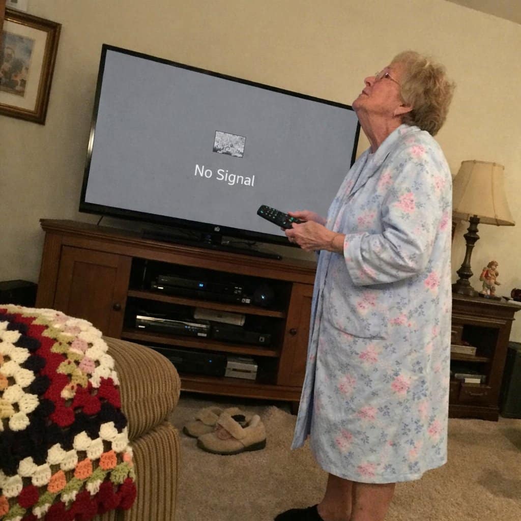 An older woman standing in front of a television pressing a remote while looking up at a gray no-signal screen, with a crocheted blanket on the sofa arm and slippers on the floor nearby