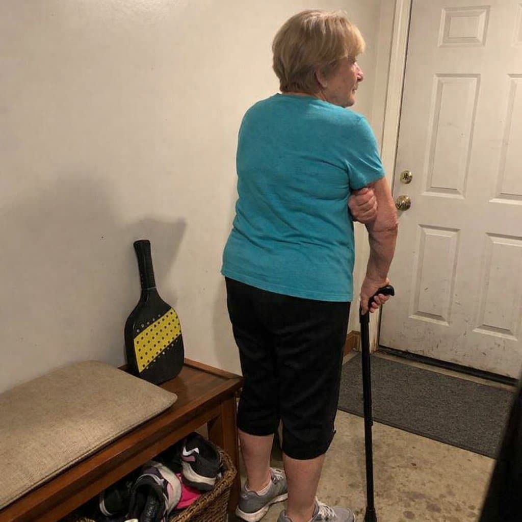 An older woman with a cane standing near a mudroom bench rubbing her elbow, a pickleball paddle resting against the wall beside her