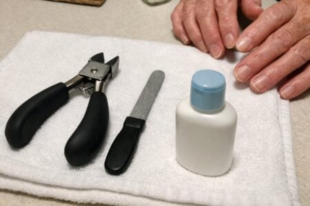 Large-handled toenail clippers and a nail file on a white towel on a kitchen table, with an older woman's hands reaching toward them