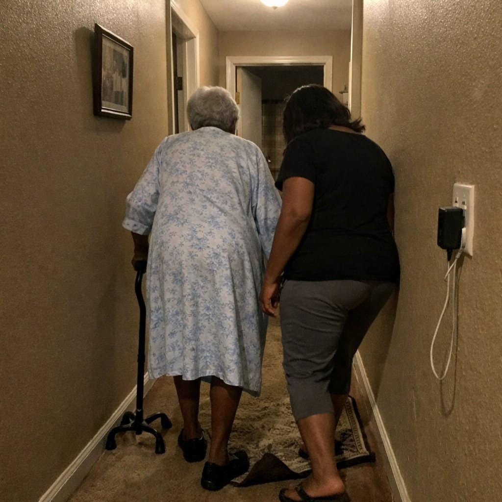 An older woman with a cane walking down a dim hallway while a younger woman beside her checks the floor and a loose rug edge