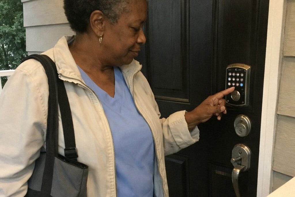 A woman in scrubs pressing a button on a door keypad at a residential front door, viewed from the side on the porch