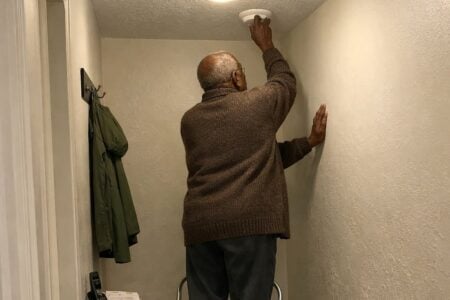 An older man in a cardigan standing on a step stool in a home hallway, reaching up with one hand braced on the wall to access a smoke alarm on the ceiling, photographed from down the hall