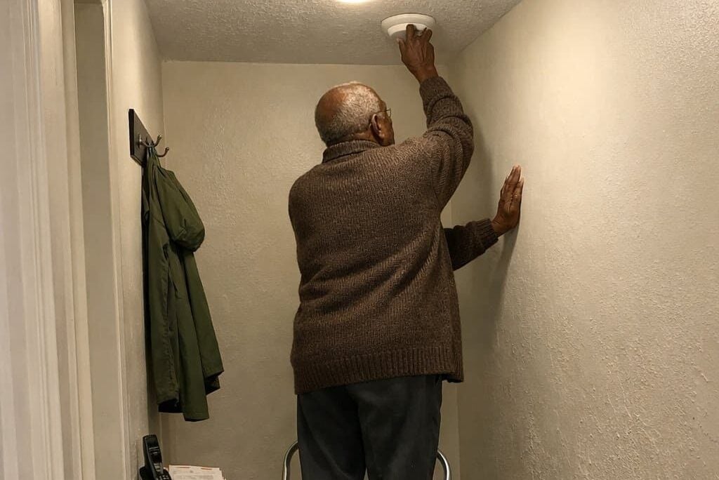An older man in a cardigan standing on a step stool in a home hallway, reaching up with one hand braced on the wall to access a smoke alarm on the ceiling, photographed from down the hall