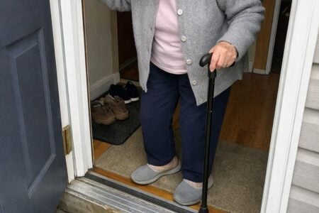 An older woman holding a cane and gripping the front door frame, looking down at the door sill before stepping out onto the porch