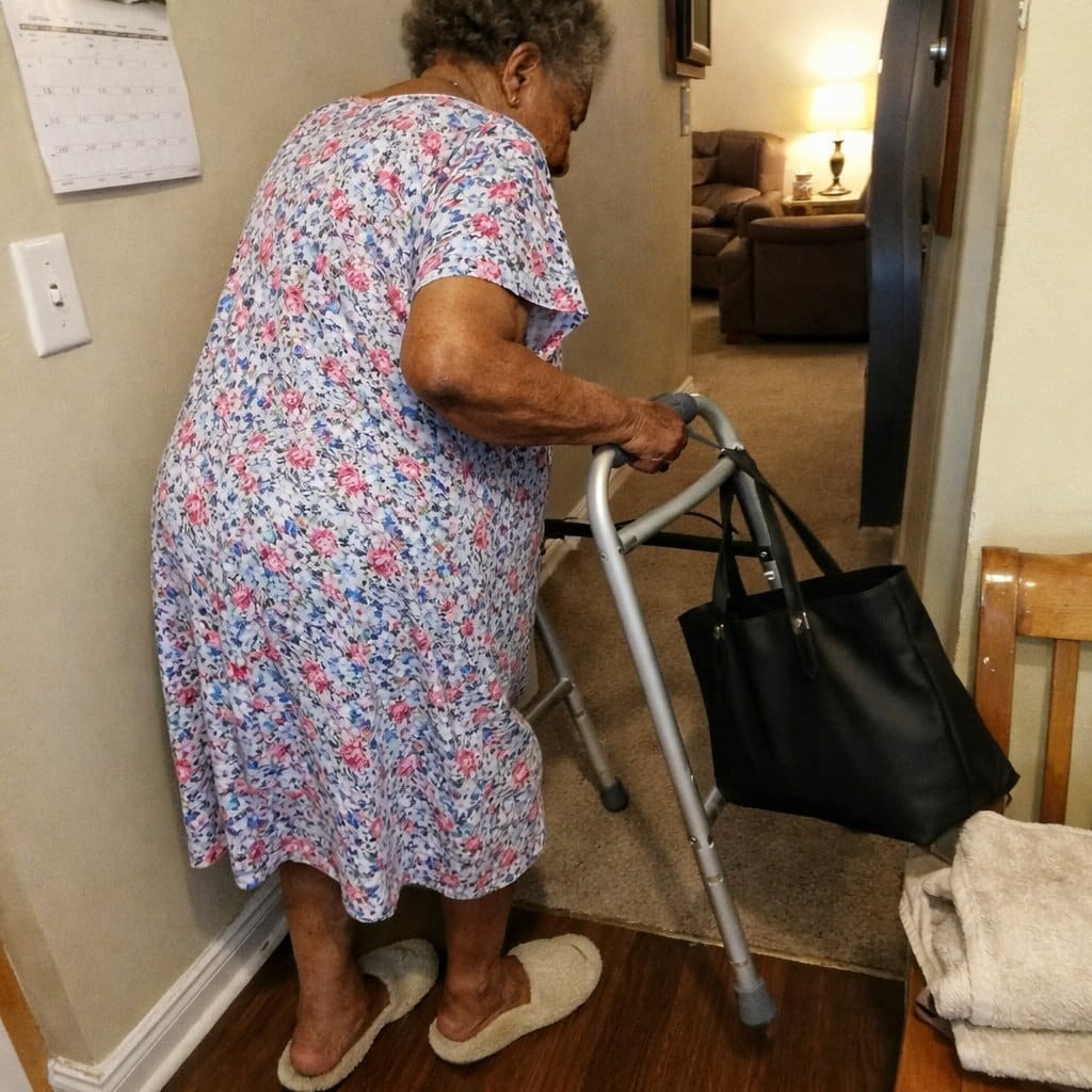 An older woman with a standard walker stopped in a hallway, the walker frame tilting to one side from an unevenly loaded bag, the woman looking down at it with a concerned expression
