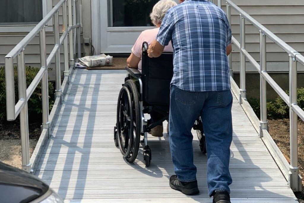 A man pushing an older woman in a manual wheelchair up a modular aluminum ramp with handrails along the side of a house toward the front door