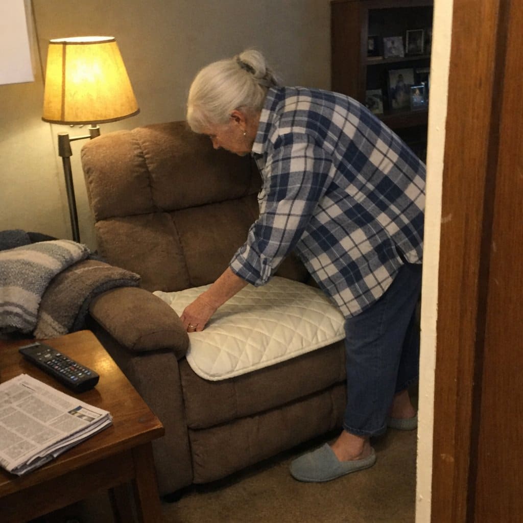 An older woman smoothing a quilted pad onto the seat of a recliner in a lived-in living room