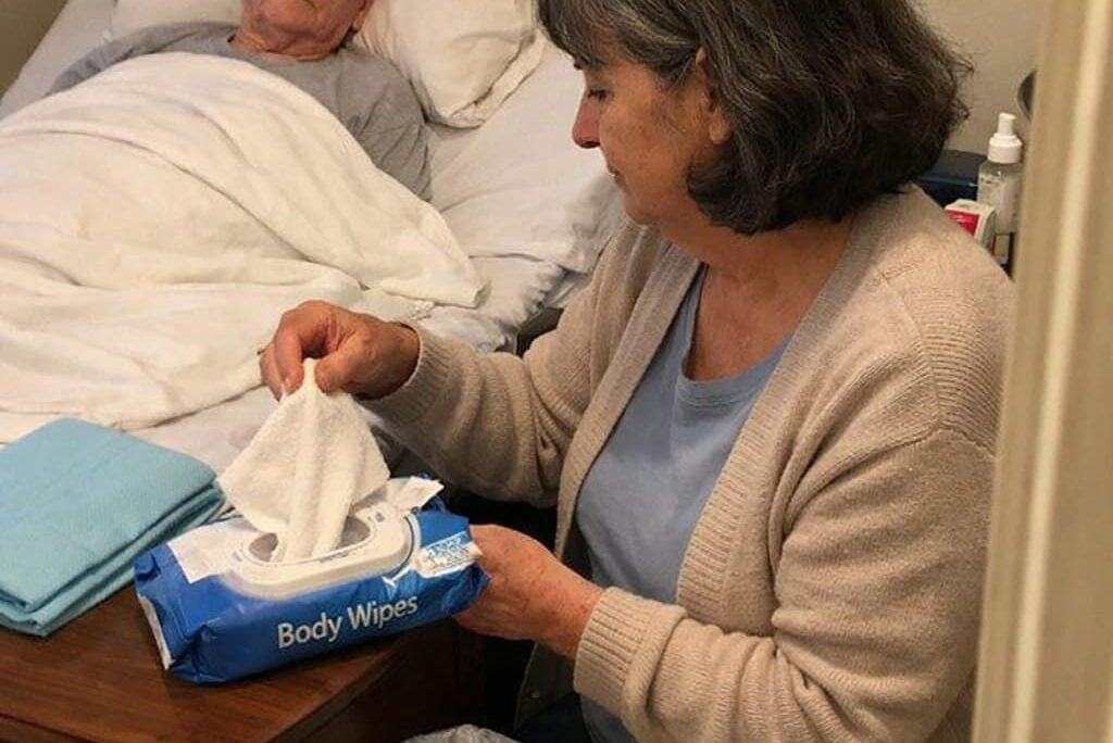 A woman pulls a large body wipe from an open package on a nightstand while an elderly man lies in the bed behind her