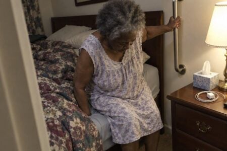 An older woman gripping a vertical bedside grab bar and pushing herself to standing at the edge of a bed, looking down at her feet, with a lit nightstand lamp beside her