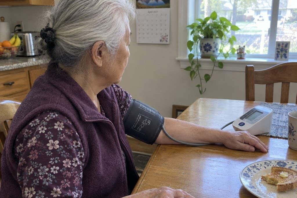 An older woman with visibly swollen knuckle joints seated at a kitchen table with a blood pressure cuff on her upper arm, free hand resting flat beside her