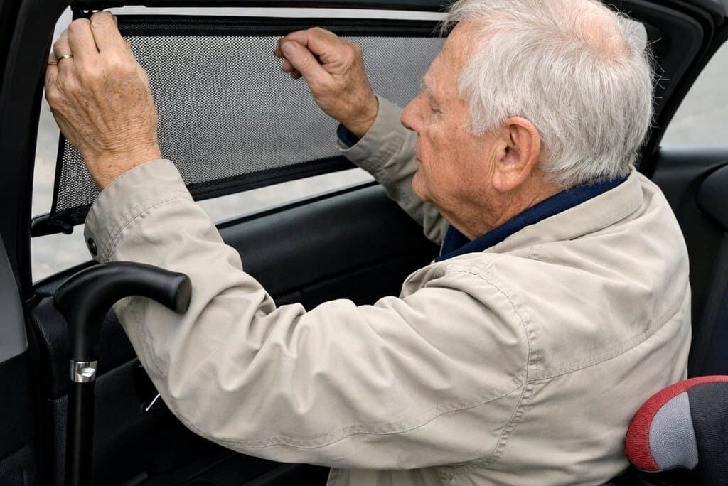 An older man with a cane hooked over a car door frame, using both hands to pull a retractable window shade across the rear passenger window