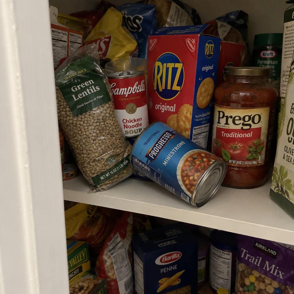 A crowded pantry shelf with cans, boxes, and food bags stacked without organization, one item tipped on its side