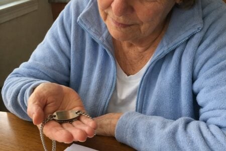 An older woman sits at a kitchen table holding a medical alert bracelet in her open palm, turning it over to examine the plaque, with a handwritten list and pen on the table in front of her