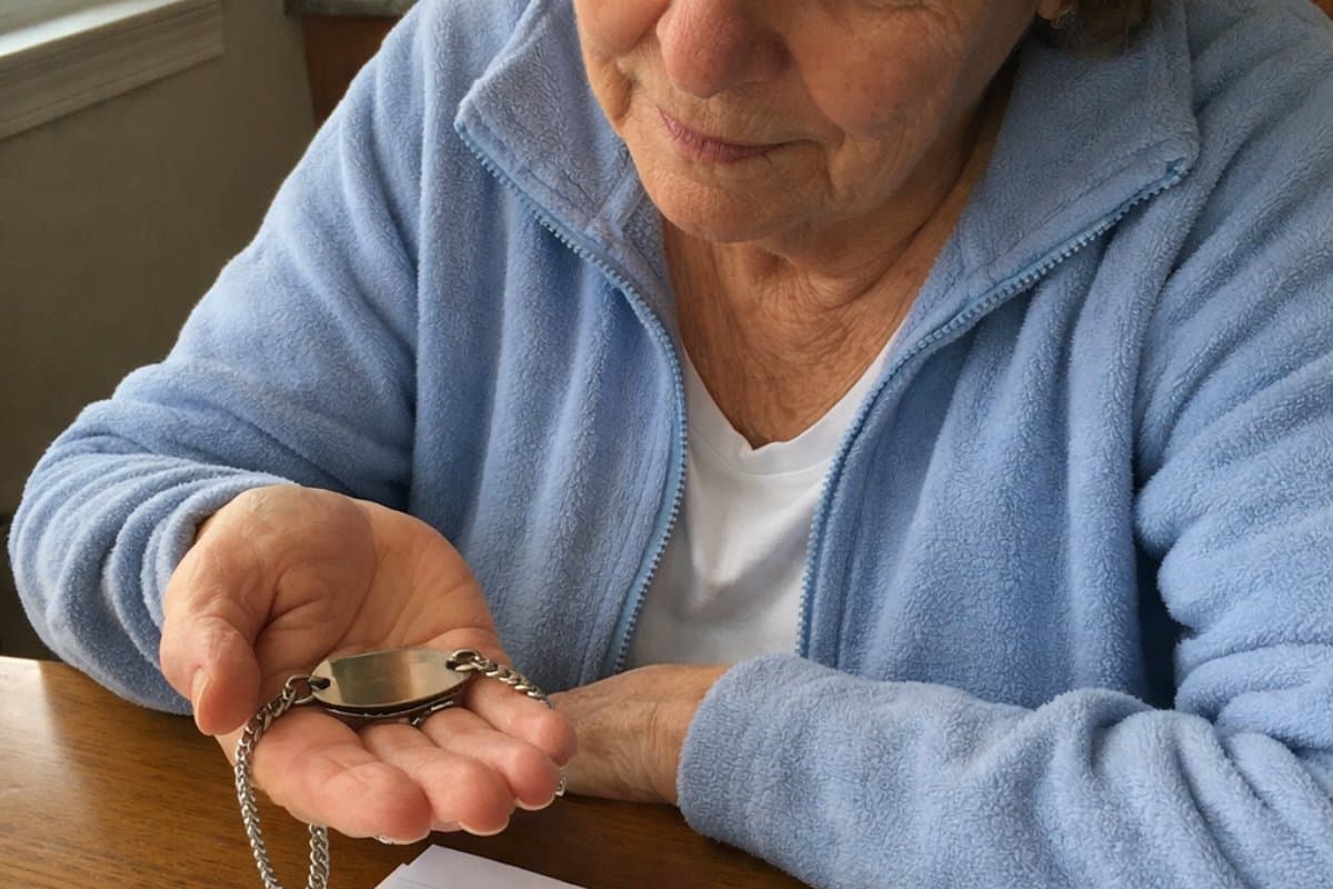 An older woman sits at a kitchen table holding a medical alert bracelet in her open palm, turning it over to examine the plaque, with a handwritten list and pen on the table in front of her