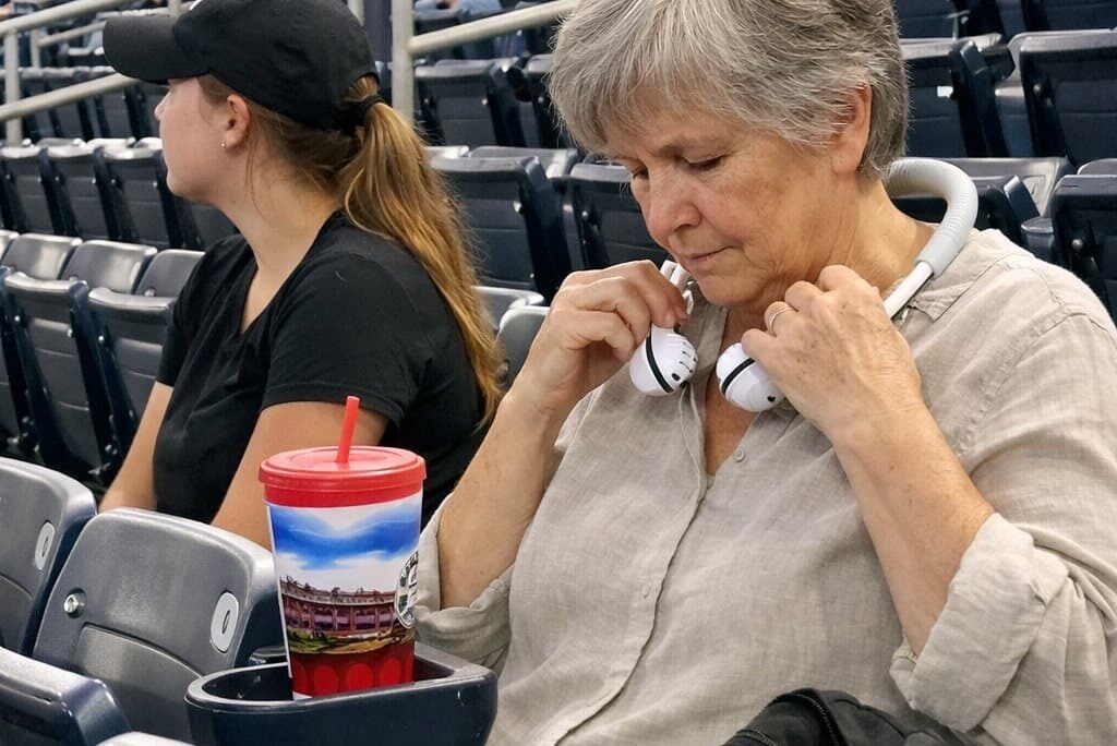 An older woman with short grey hair sits on bleachers fastening a neck fan around her neck while a young adult beside her watches the field