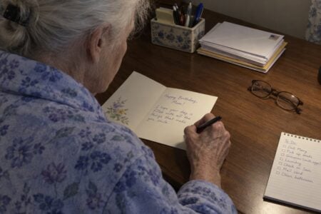 An older woman sitting at a home writing desk, looking down at a birthday card she is writing, with envelopes, a notepad, and reading glasses on the surface beside her