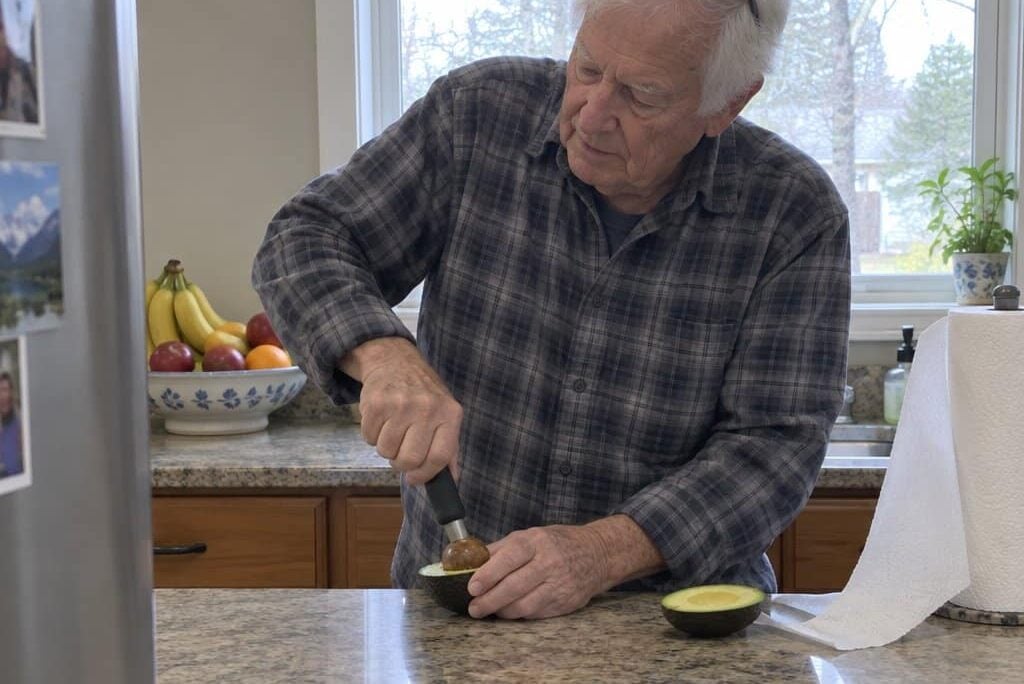 An older man pressing an avocado half on a counter and working out the pit with a kitchen tool, slightly turned toward the task