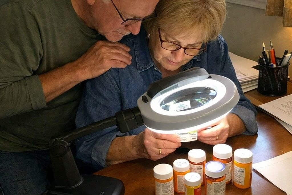 An older man and woman bent together over a lit desktop magnifier on a home office desk, both looking through it at a prescription label, with several other medication bottles arranged nearby