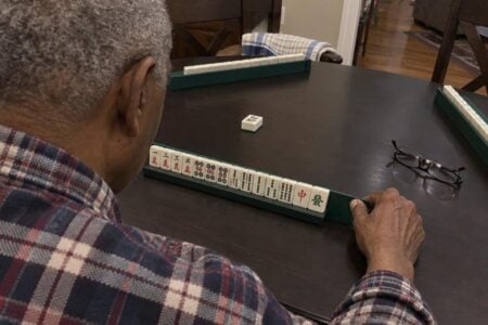 An older man with grey hair reaches forward to press mahjong tiles against a loaded tile rack at a dining table, with additional racks visible at other seats