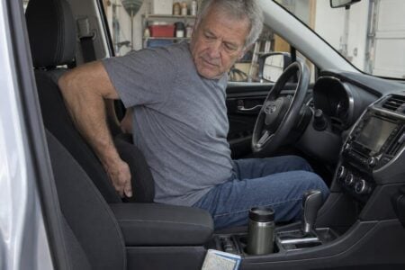 An older man sitting in a parked car driver's seat, reaching behind him with both hands to reposition a lumbar pillow before driving