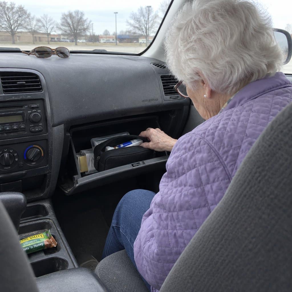 An older woman in the passenger seat looks through a small zippered kit resting in an open glove compartment