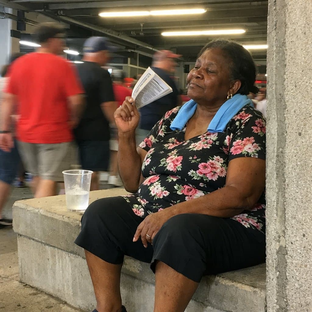 An older woman sits on a low wall in stadium shade with a cooling towel on her neck and a paper program in her hand, eyes closed