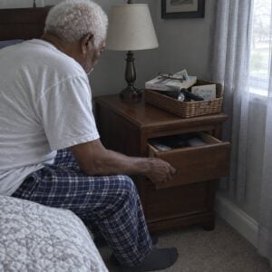 An older man sitting on the edge of a bed pulls open a nightstand drawer, with a small basket holding a flashlight visible on top of the nightstand