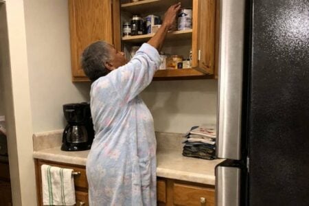 An older woman stands in her kitchen with her arm stretched upward toward the top shelf of an open cabinet, her weight shifted back on her heels as she reaches