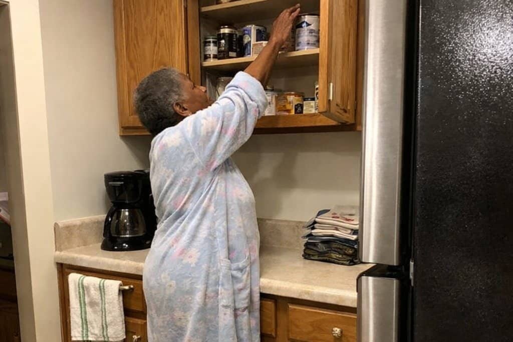 An older woman stands in her kitchen with her arm stretched upward toward the top shelf of an open cabinet, her weight shifted back on her heels as she reaches