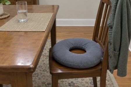 An open-center donut cushion placed slightly off-center on a wooden dining chair pulled away from a table, the round opening visible, a cardigan draped over the chair back