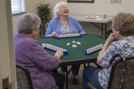Three older women, one with a cane on her chair, laugh and play mahjong at a card table in a community common room