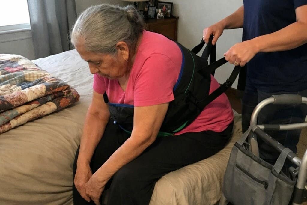 An older woman seated at the edge of a bed wearing a transfer sling and leaning forward while a caregiver standing to her side holds both sling handles