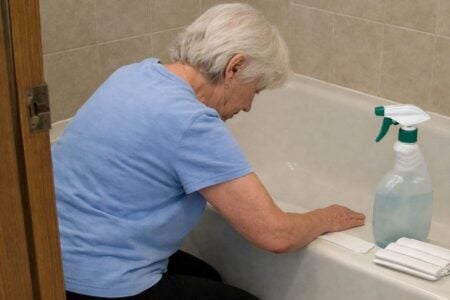 An older woman kneeling beside a bathtub pressing an adhesive strip tread flat against the tub floor with her hand, her head turned down watching what she is doing
